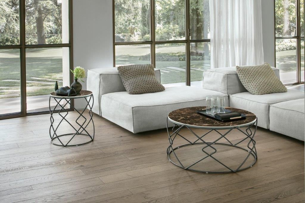 Soggiorno con divano bianco, due tavoli con struttura in metallo e ampie finestre. Pavimenti marroni e pareti chiare.