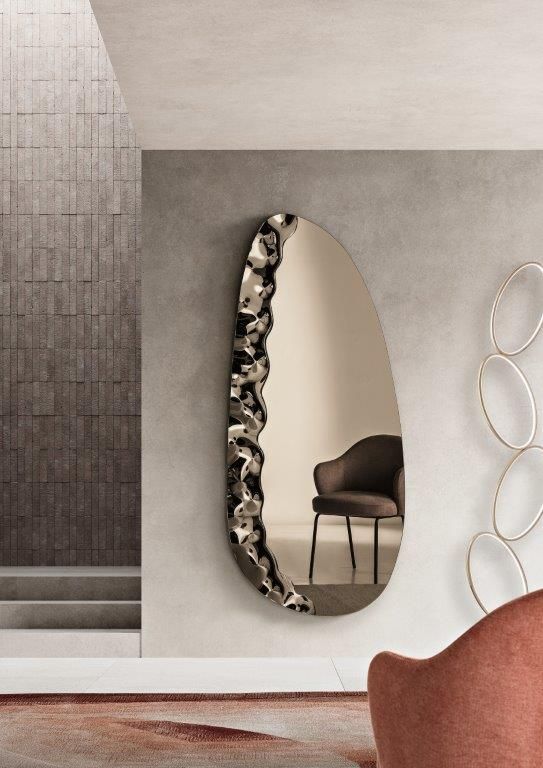 Specchio ovale con cornice metallica testurizzata, appeso a una parete grigia. Interno con sedia e decorazioni murali.