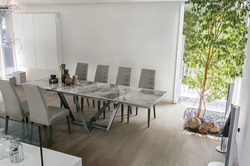 Sala da pranzo con tavolo in marmo, sedie grigie e albero decorativo vicino a una finestra.