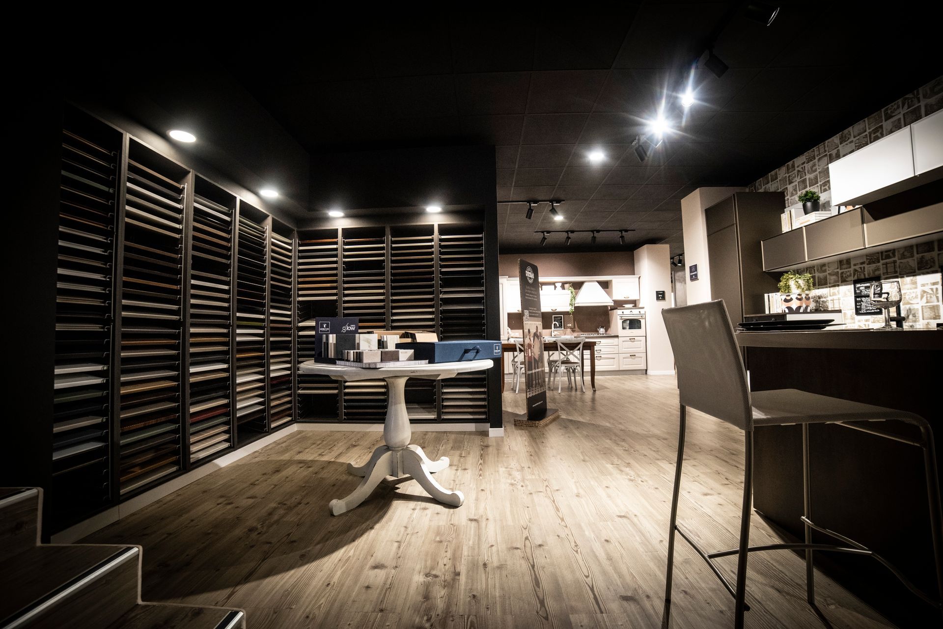 Interno di uno showroom di cucine con esposizione di campioni, tavolo, sgabelli da bar e set da cucina.