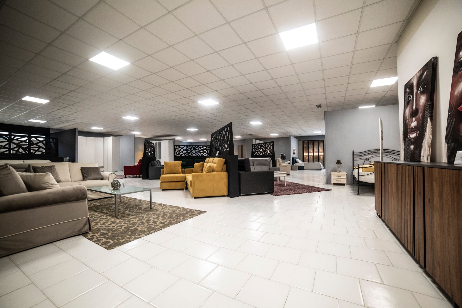 Ampio showroom con esposizione di mobili. Divani beige, poltrone gialle, opere d'arte e pavimento piastrellato.