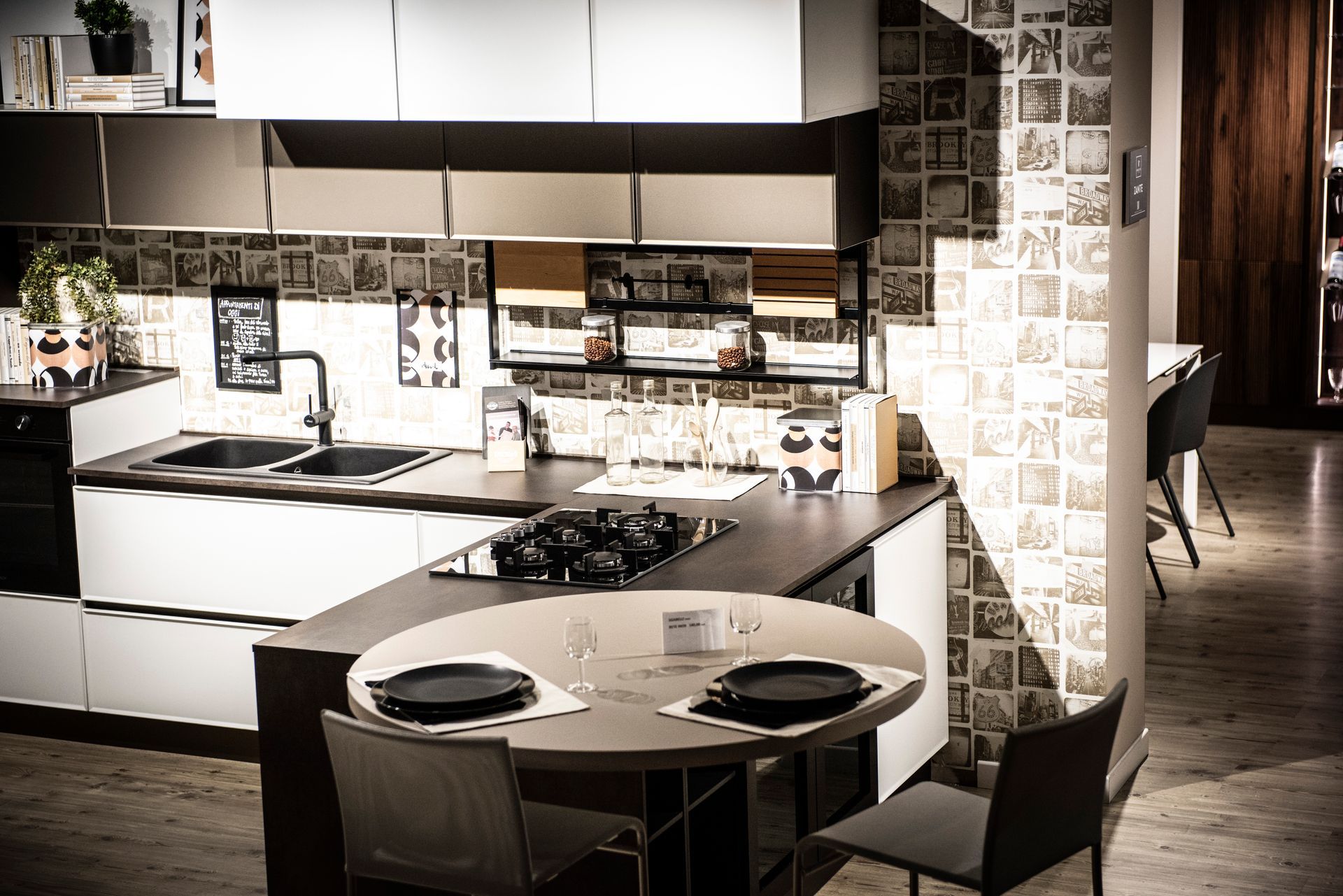 Cucina moderna con mobili bianchi e grigi, ripiani neri e un piccolo tavolo rotondo con due sedie.