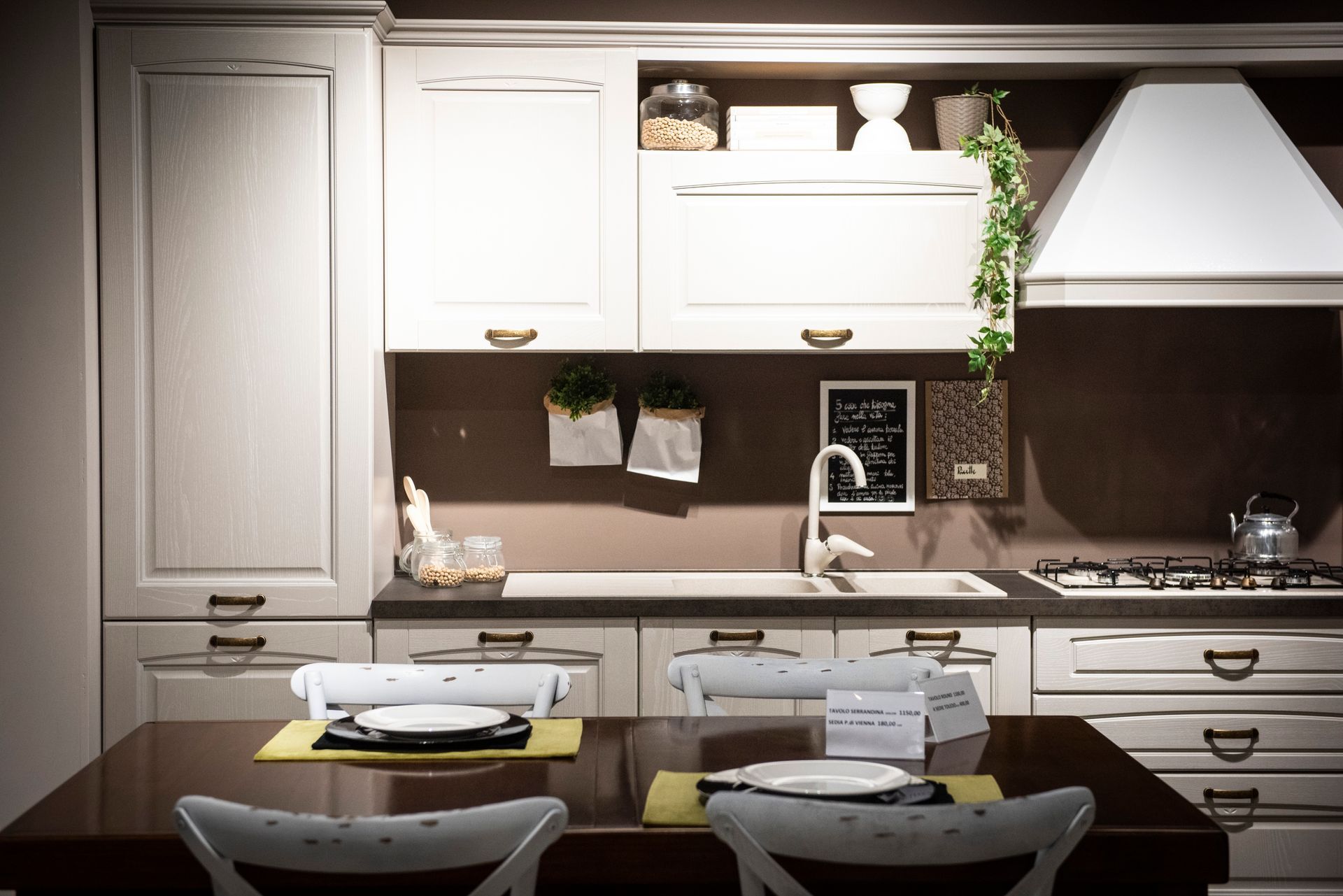 Mobili da cucina color crema con piano di lavoro e tavolo da pranzo scuri; oggetti decorativi sulla parete e sul tavolo.