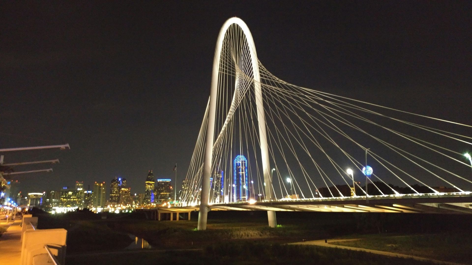 Night view of the Margaret Hunt Hill Bridge in Dallas, lit with white lights, with city skyline in the background.