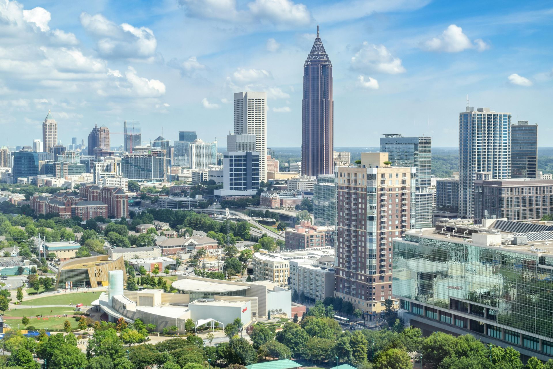 Atlanta skyline with skyscrapers under a blue sky, viewed from a high angle, with green trees in the foreground.