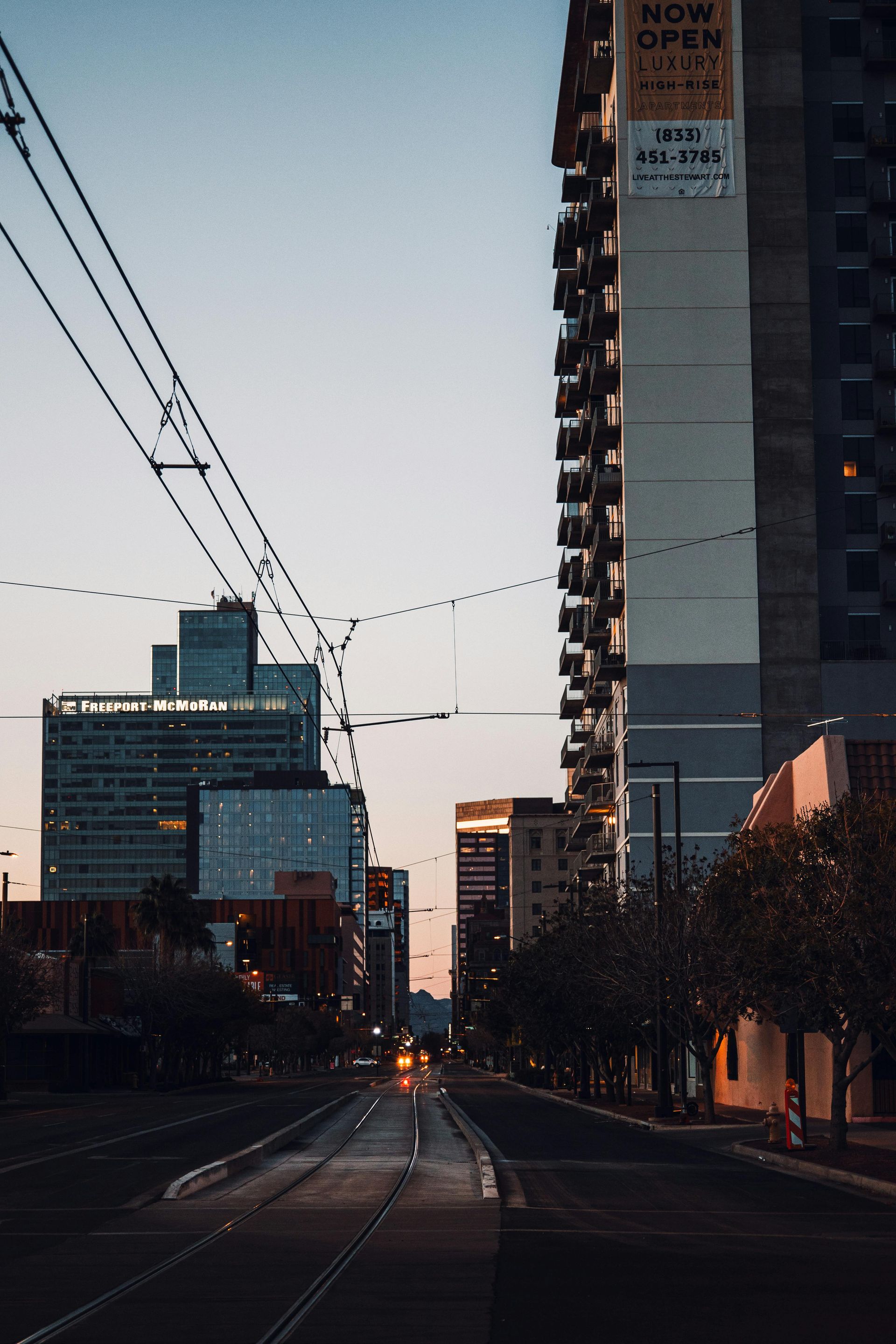 City street scene at dusk with buildings, trolley tracks, and overhead wires.