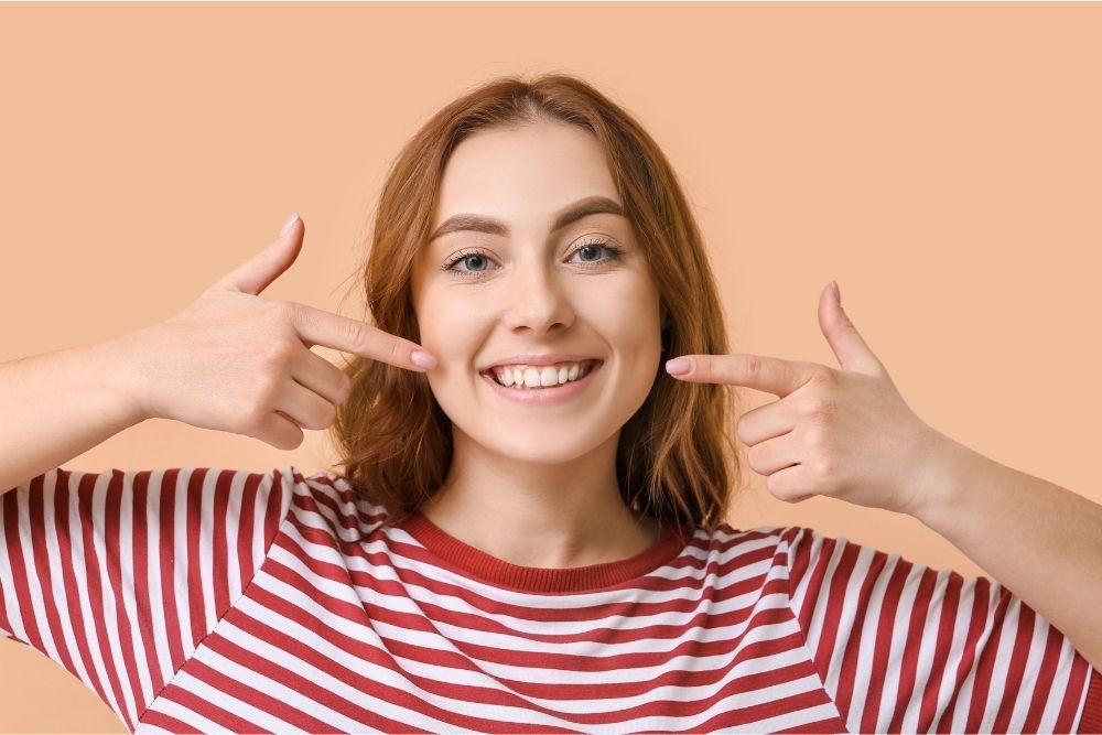 Woman pointing at her smile, wearing a striped shirt, against a peach background.