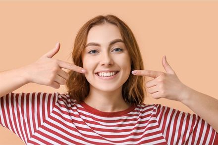 Woman pointing at her smile, wearing a striped shirt, against a peach background.