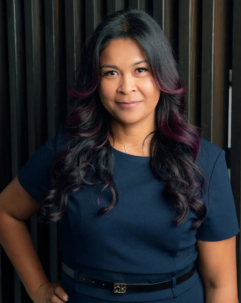 Woman in a navy dress with long dark hair, purple highlights, and a slight smile. Against a dark background.