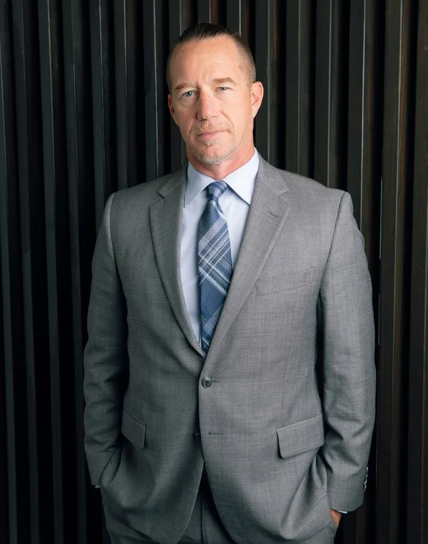 Man in gray suit with hands in pockets, standing in front of a dark, textured wall.