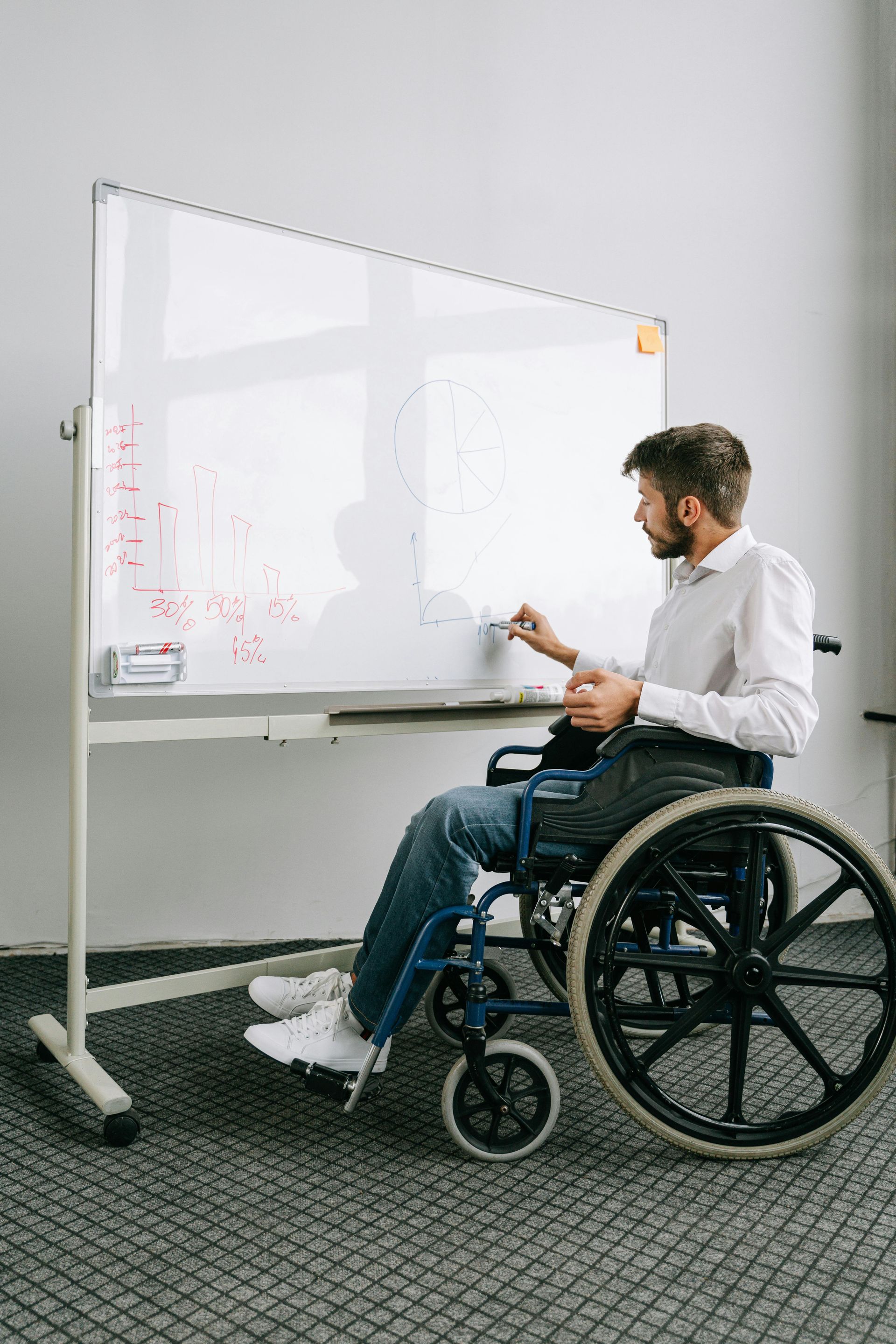 Man in wheelchair writing on a whiteboard in a bright office.