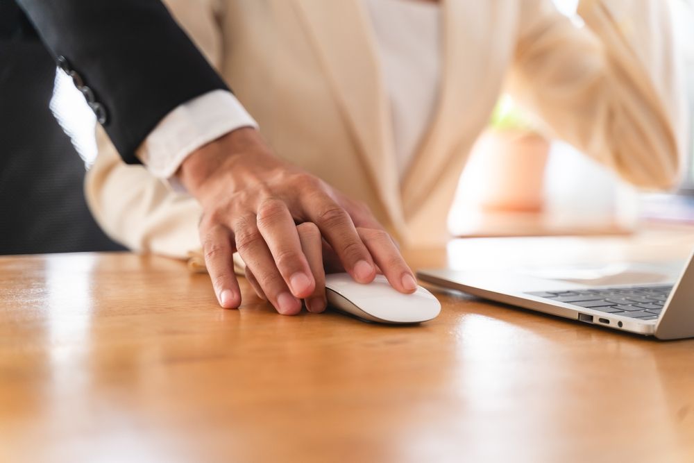 Person's hand on a computer mouse, another person's hand on top of it near a laptop on a wooden desk.