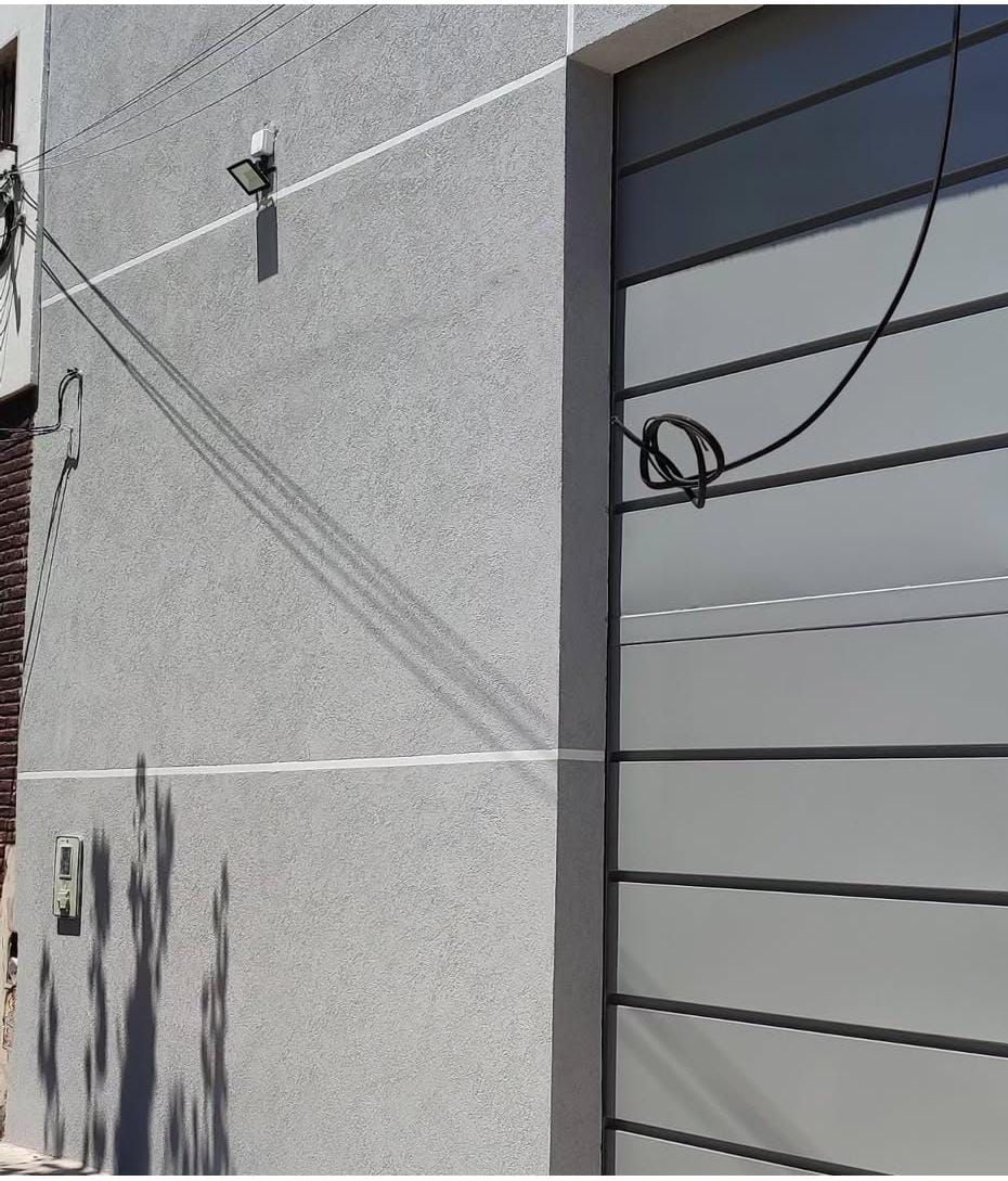 Fachada del edificio de color gris texturizado con luminaria y puerta de garaje gris claro. Cable negro colgando.