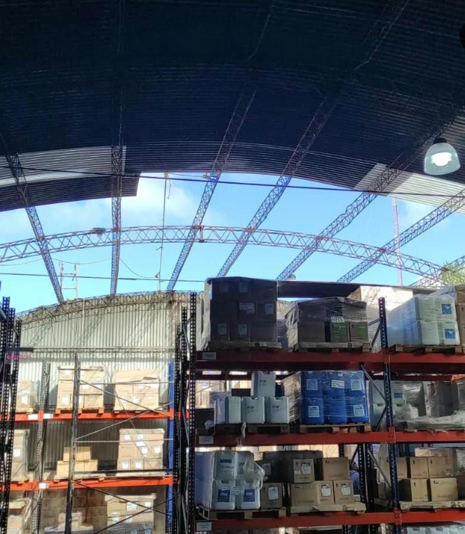 Interior de almacén con estanterías repletas de mercancía, red en el techo y cielo visible.