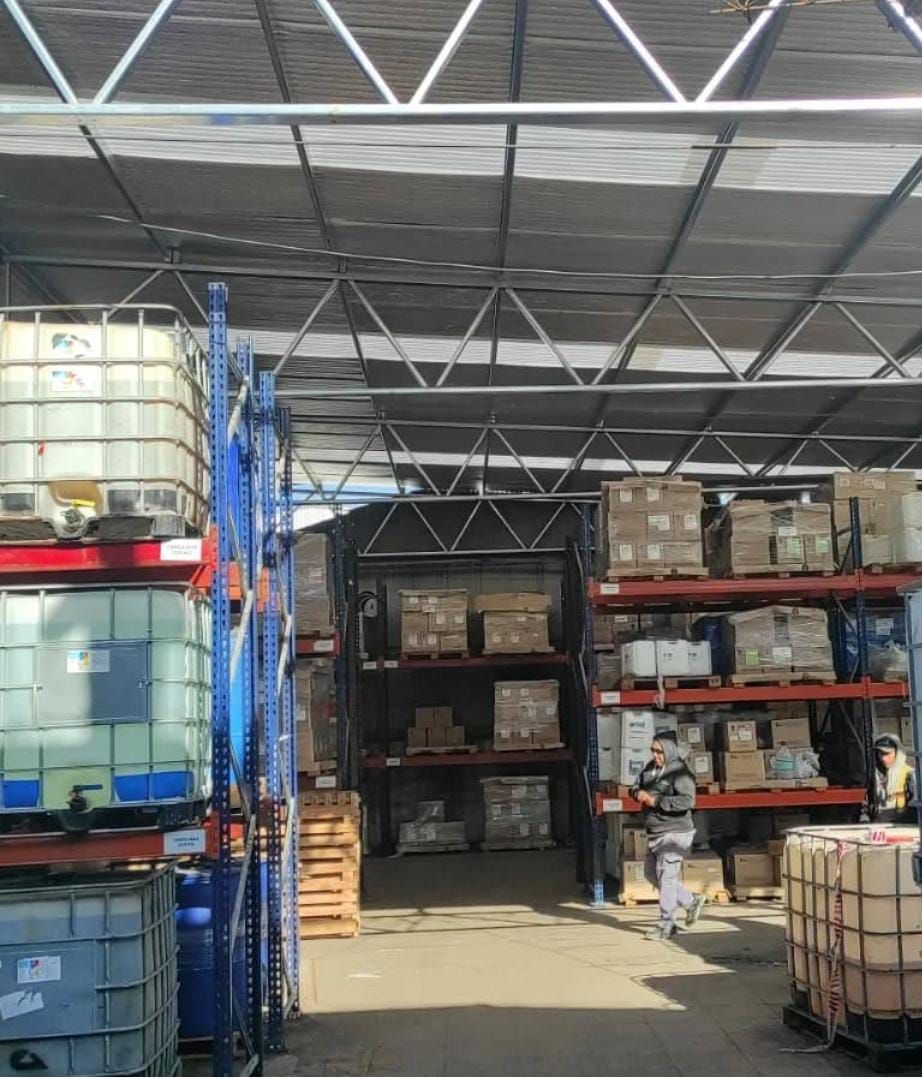 Interior de almacén con mercancía apilada en estanterías. Techo metálico, estanterías azules y presencia de personas.