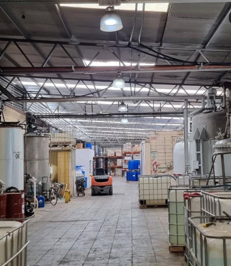 Interior de almacén con montacargas, tanques de almacenamiento y contenedores. Se ven luces superiores y vigas metálicas.