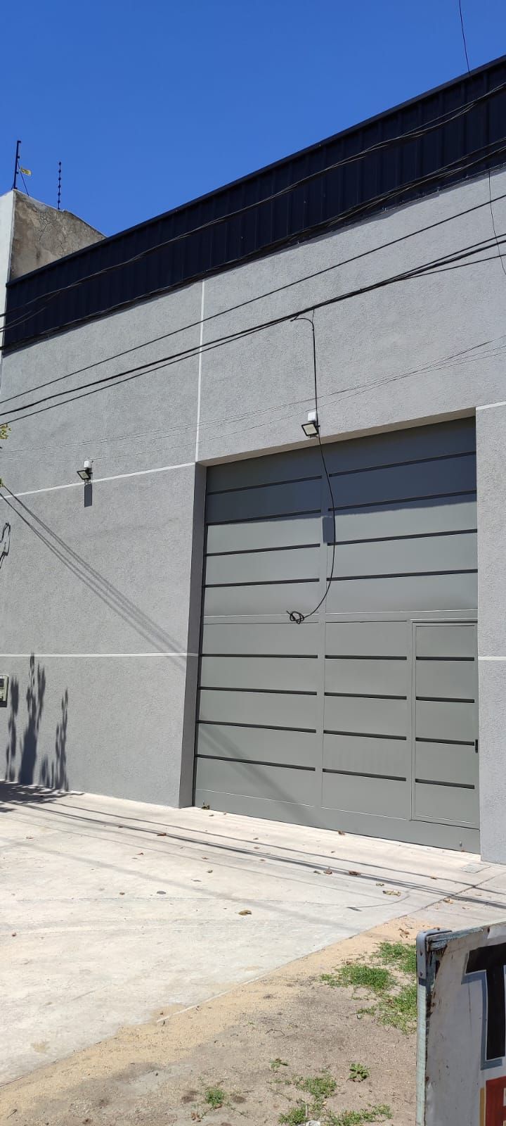 Edificio industrial gris con puerta de garaje cerrada y cables de electricidad contra un cielo azul.