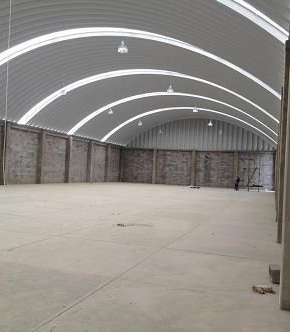 Interior vacío de un almacén con suelo de hormigón, paredes grises y techo metálico arqueado con claraboyas.