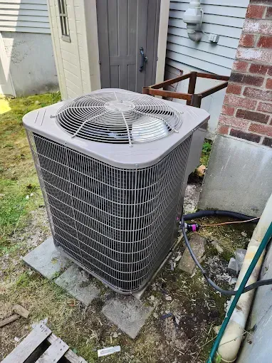 A gray air conditioning unit outside, next to a shed and brick wall. The unit is on concrete slabs, with a hose and pipes visible.