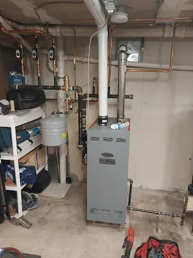 A gray furnace in a basement with copper pipes and a white vent. A cluttered shelf and tools are in the foreground.