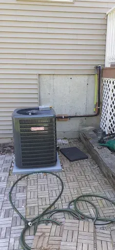 An air conditioning unit on a patio near a house. A green hose is coiled on the pavers.
