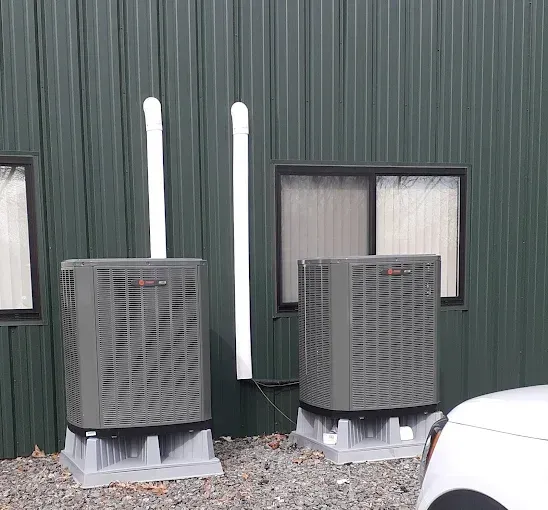 Two gray HVAC units with white vent pipes outside a green building, next to a car.