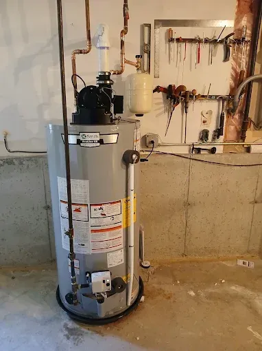 A gray water heater in a basement with copper pipes, a white expansion tank, and a wall with tools.
