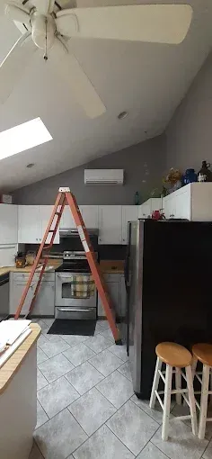 A kitchen with a ladder leaning against a wall; an air conditioner is mounted high on the wall. A fridge and stools are in the right foreground.