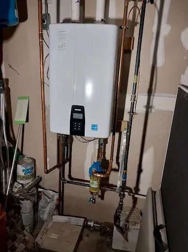 A wall-mounted, white tankless water heater with copper piping in a garage setting. It has a digital display and various valves and gauges below.
