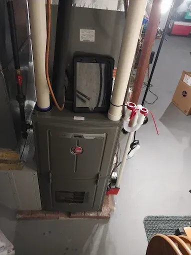 A Rheem furnace in a basement setting. Two white PVC pipes are attached, and a tablet rests on top.