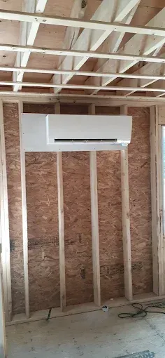 White air conditioning unit installed in a room under construction; wooden studs and ceiling beams are visible.