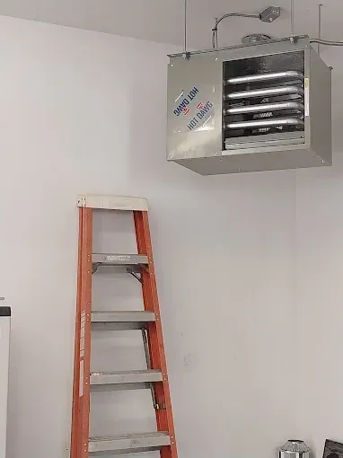 A wall-mounted electric heater in a garage, with a ladder in front of it. The heater is silver, and the ladder is orange.