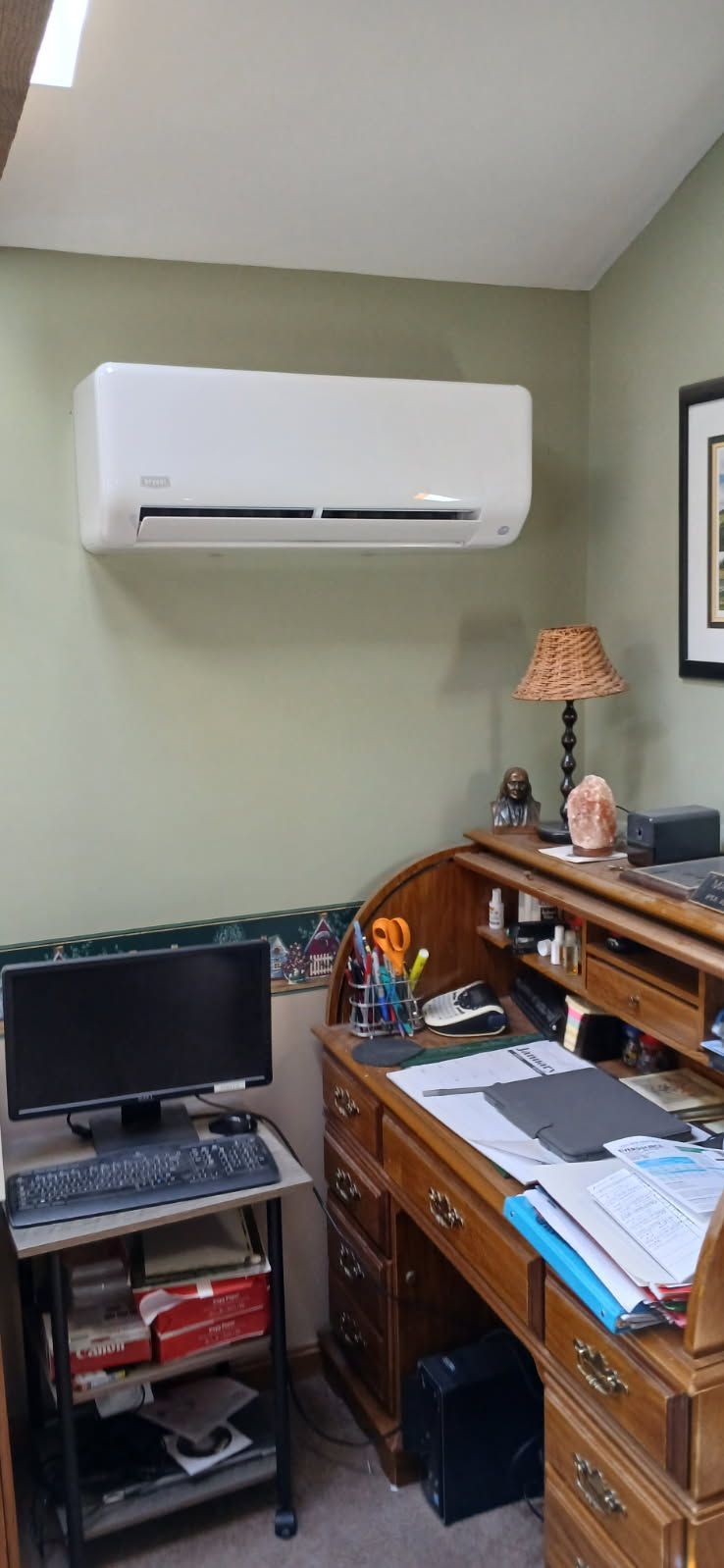 A wall-mounted air conditioning unit above a wooden desk in an office. A computer monitor, lamp, and various office supplies are visible.