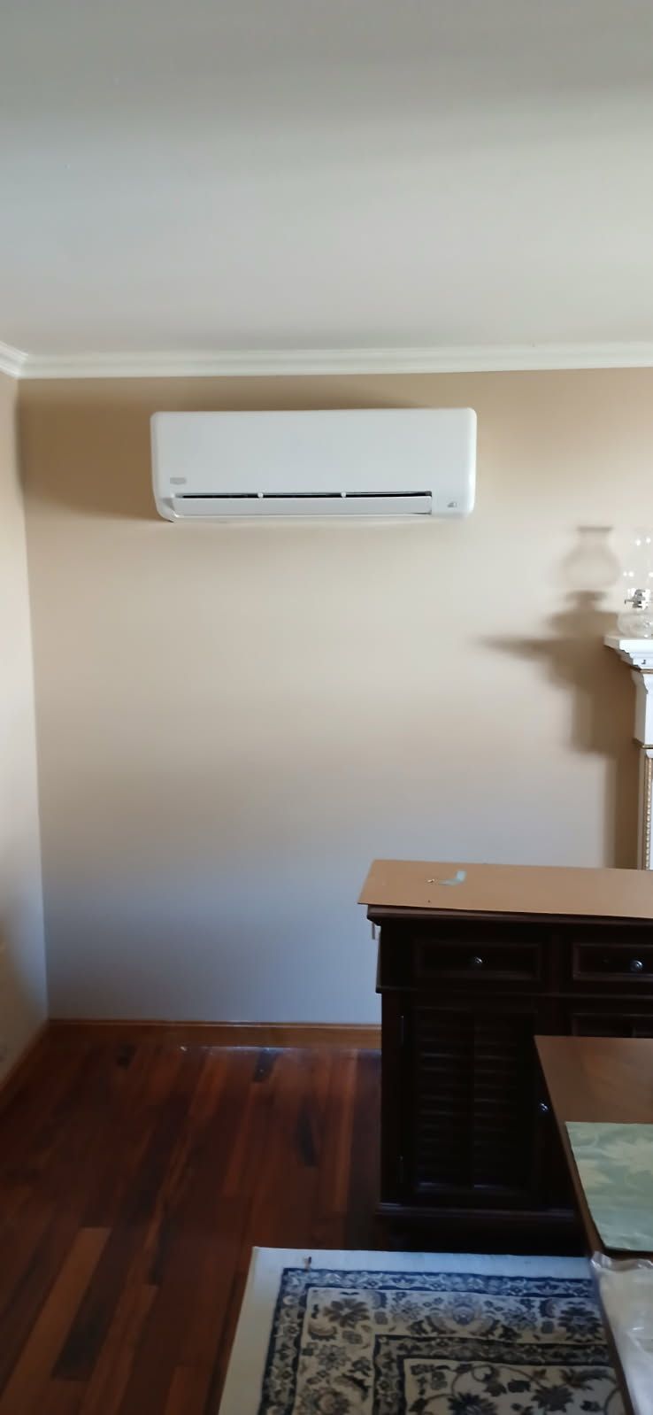 An air conditioning unit is mounted on a beige wall. Dark wooden furniture and flooring fill the room.