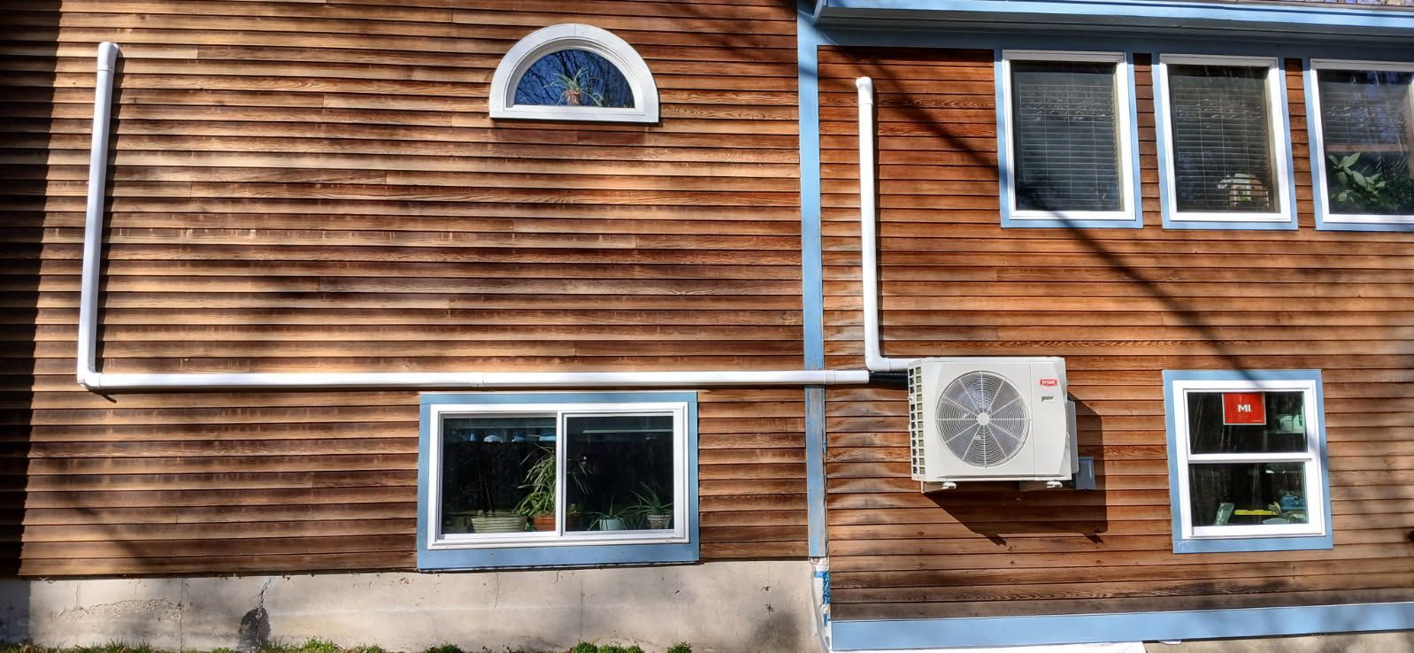 Exterior of a wooden building with a white pipe running down the wall, an air conditioning unit, and windows.