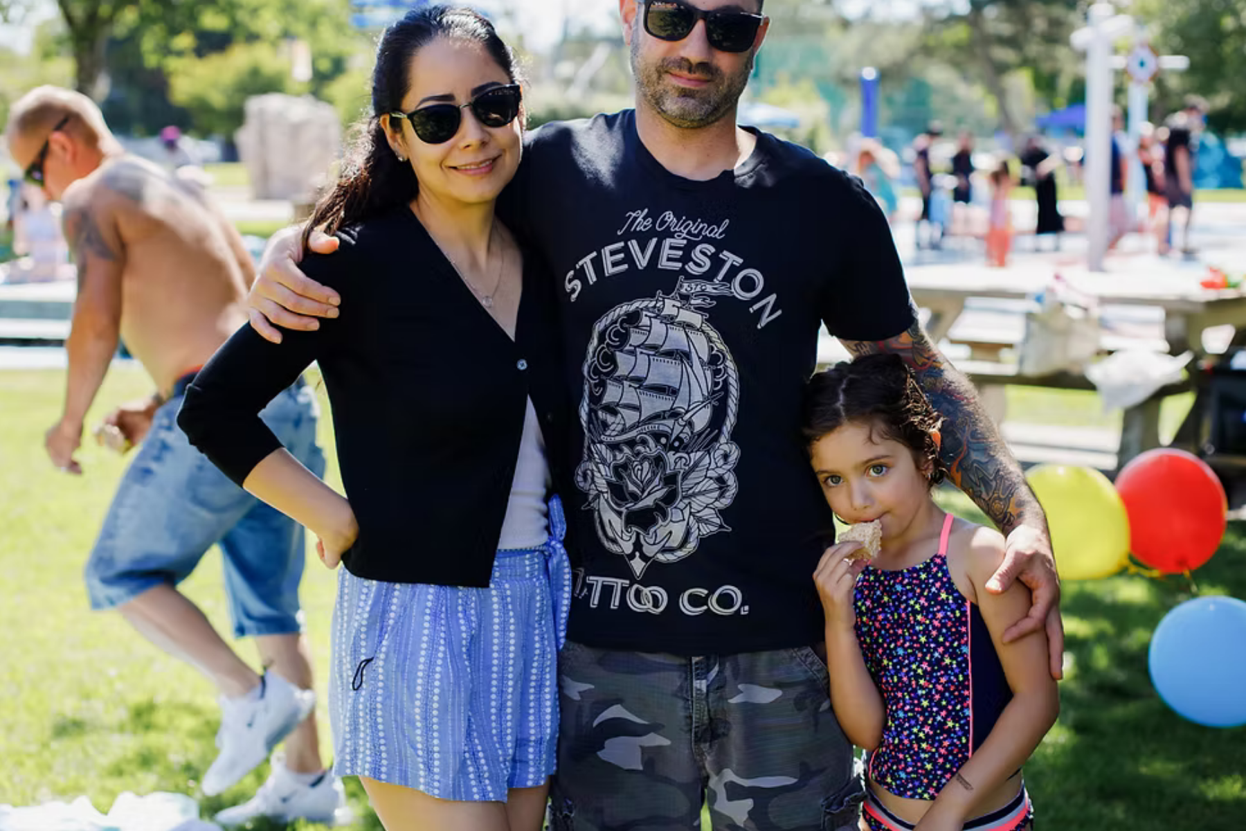 Family of three poses outdoors; woman, man in sunglasses with tattoo, and child holding treat, park background.