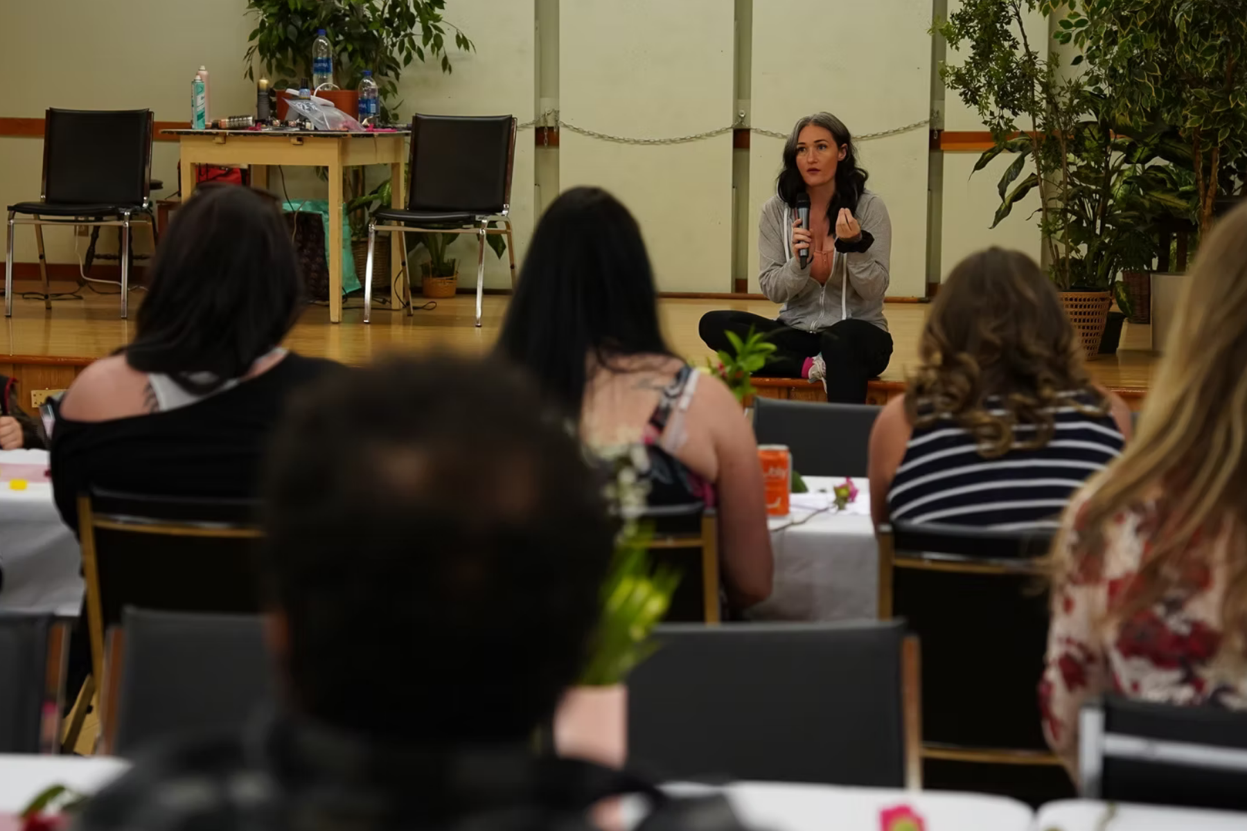 Woman speaking on stage to an audience seated at tables. Tables are decorated with flowers.
