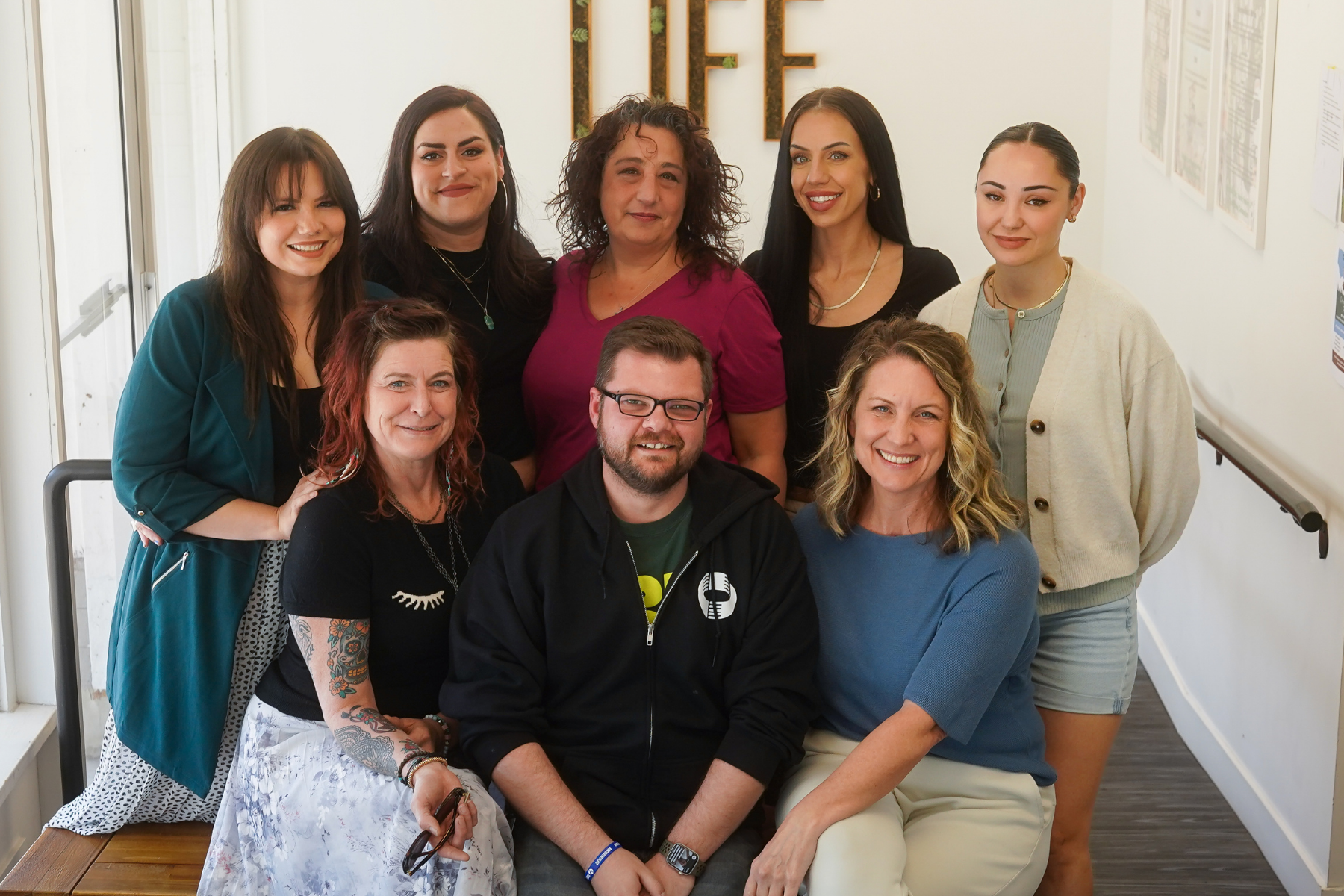 Group of eight people posing, smiling, and looking at the camera indoors, in front of a white wall.