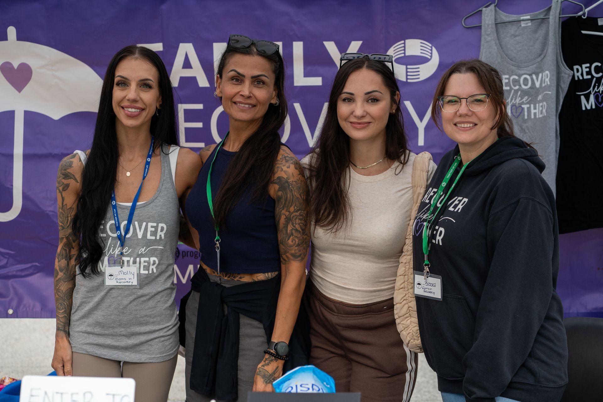 Four people smiling, posing together in front of a “Family Recovery” banner with a purple umbrella and heart graphic.