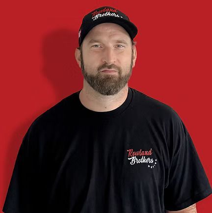 Man wearing a black hat and t-shirt with a logo, standing in front of a red background.