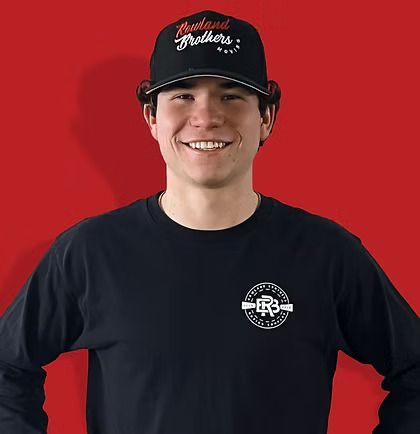 Man smiling, wearing a black cap and long-sleeved shirt, standing in front of a red background.