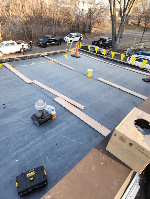 Roofing work in progress on a flat roof with tools, materials, and safety barriers near parked cars.