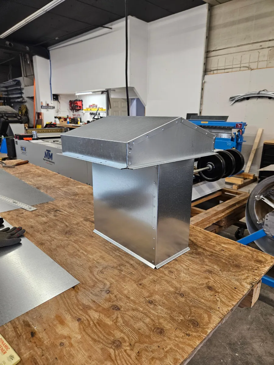 Metal HVAC duct hood on a workbench in a workshop, with tools and machinery in the background