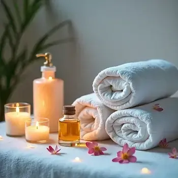 Spa towels, candles, and oil bottles arranged with pink flowers on a soft white surface.