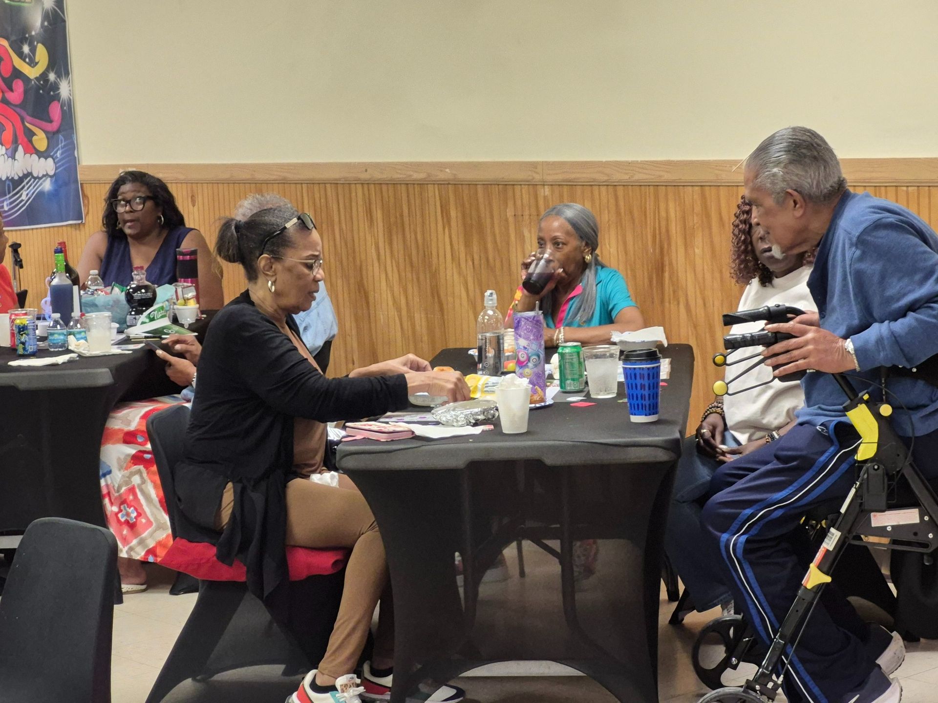 People at tables, one with walker, in a room. Someone hands something to another. Drinks and decorations are present.
