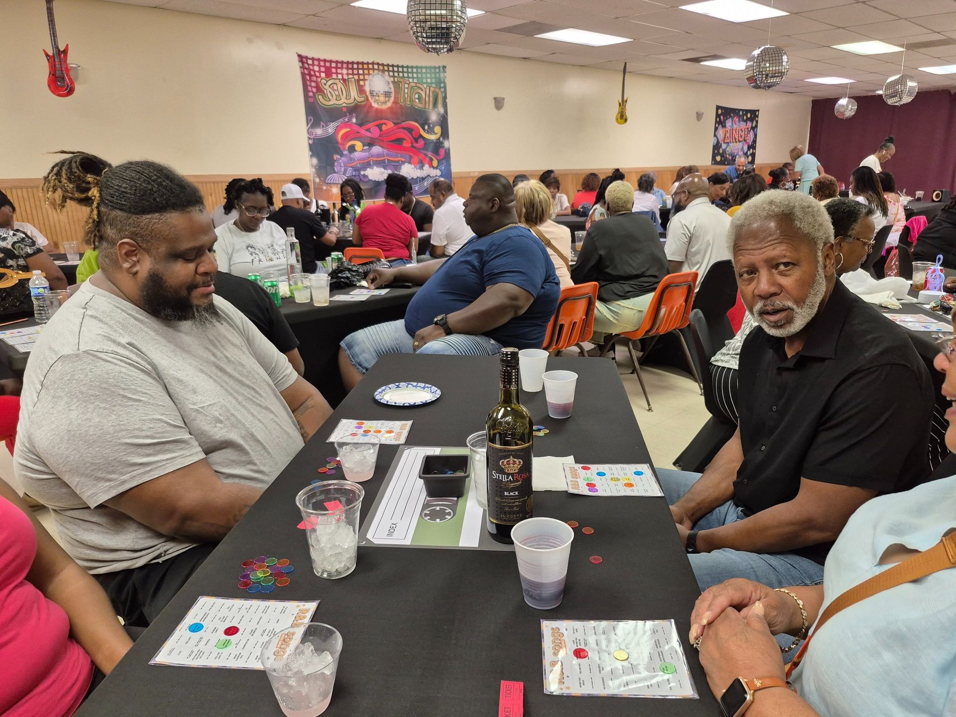 People at tables in a community hall play a game, likely bingo. Drinks and cards are visible.