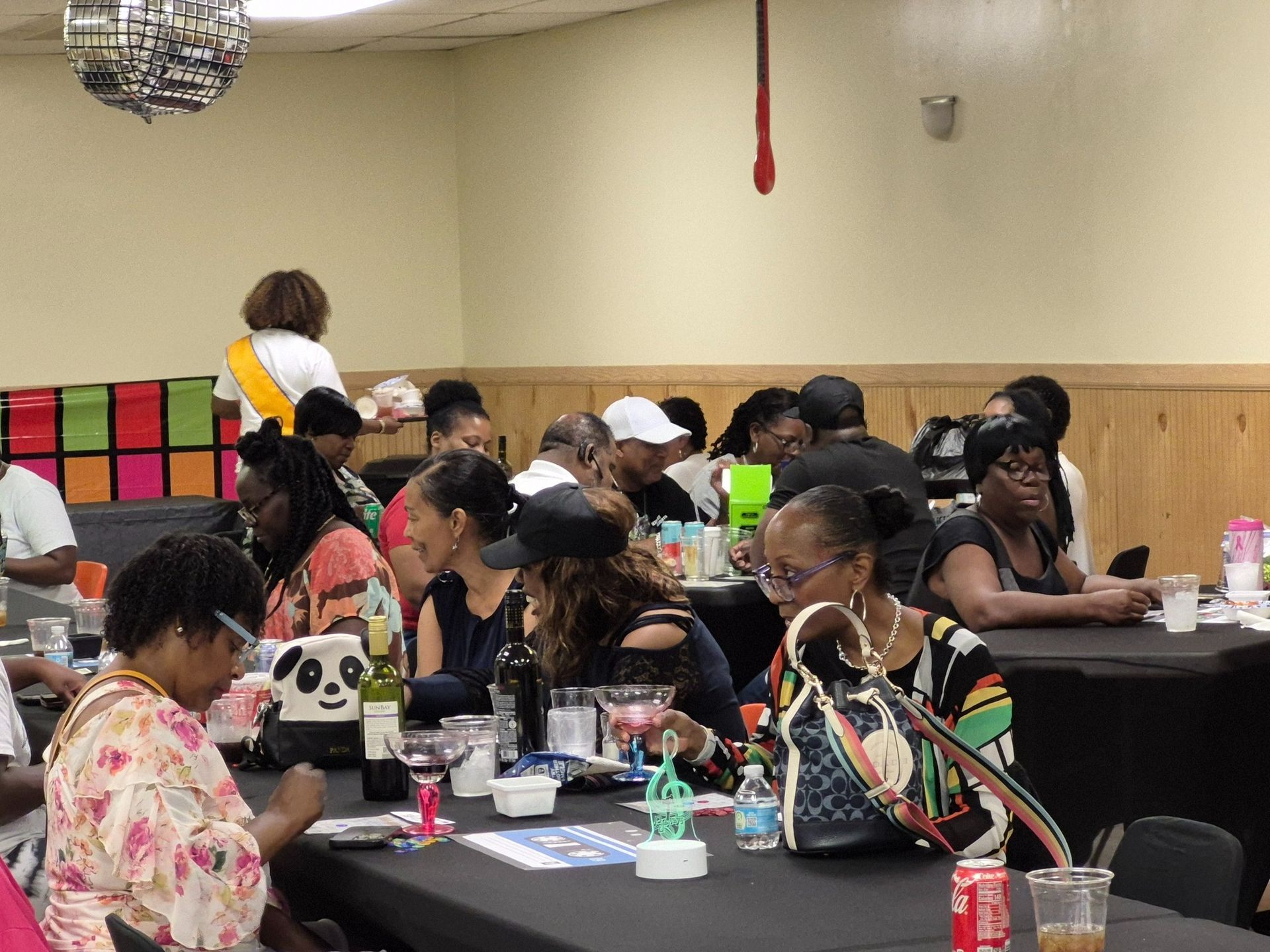 People at tables in a brightly lit room, socializing. Some people are holding drinks.