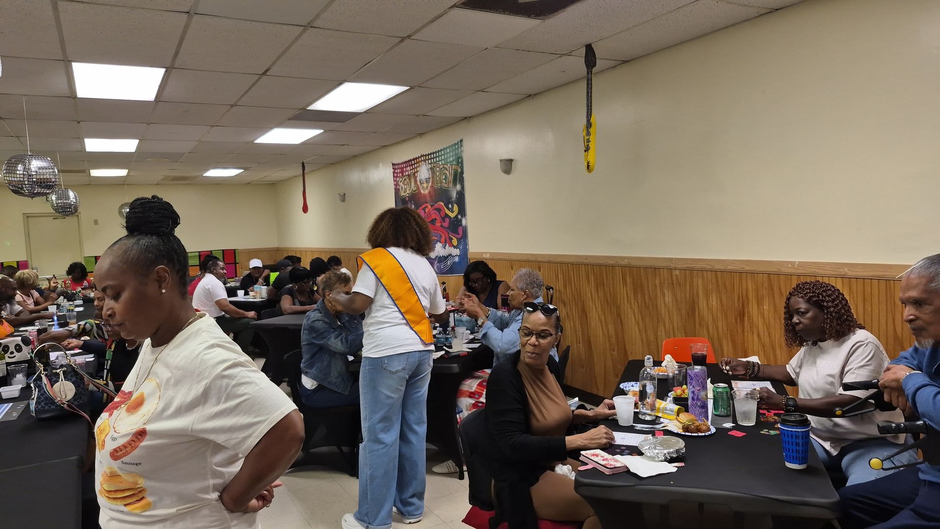 People seated at tables in a community hall. One woman wearing a sash stands near tables.