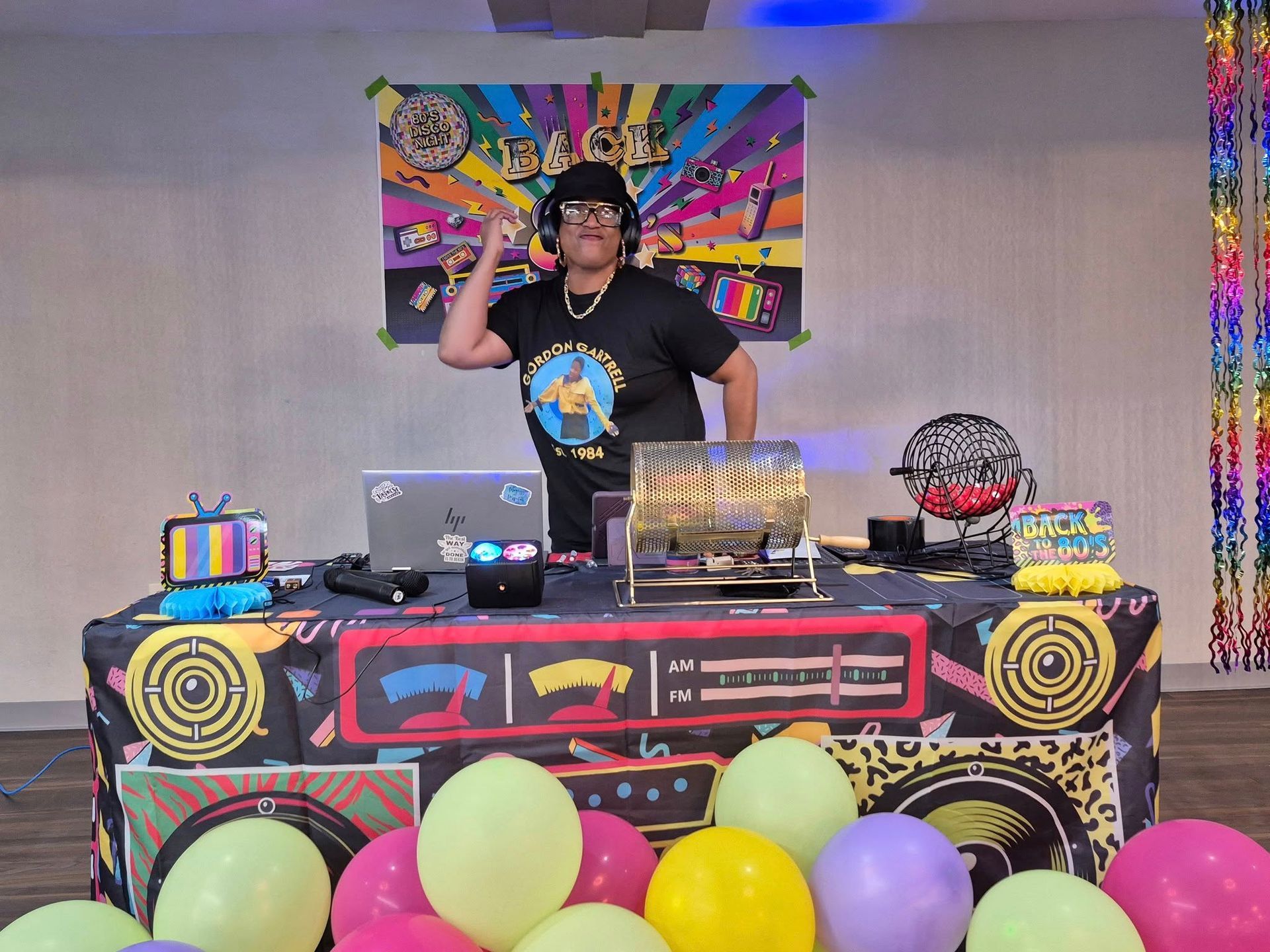 DJ at a colorful party, throwing a fist up in the air behind a DJ setup covered in festive decorations.