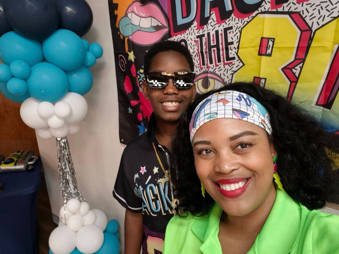 Woman and child smiling, posed next to balloons and a party backdrop.